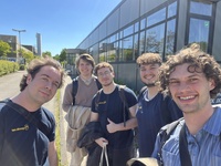 Five young men smiling for a selfie in front of a modern glass office building.